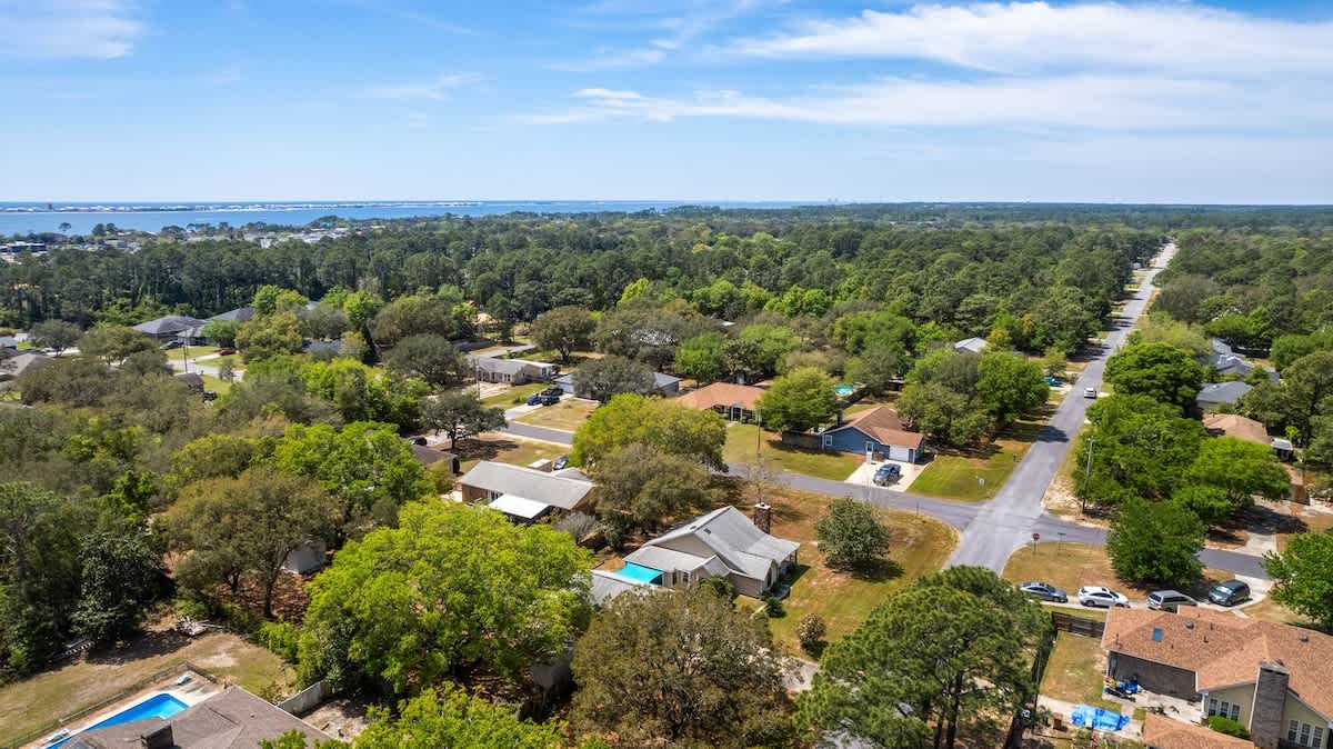 A wide aerial view showcasing the extensive tree cover of the neighborhood, with the property nestled among the greenery. The vast expanse of water and distant landmasses are prominent on the horizon, indicating a coastal location.