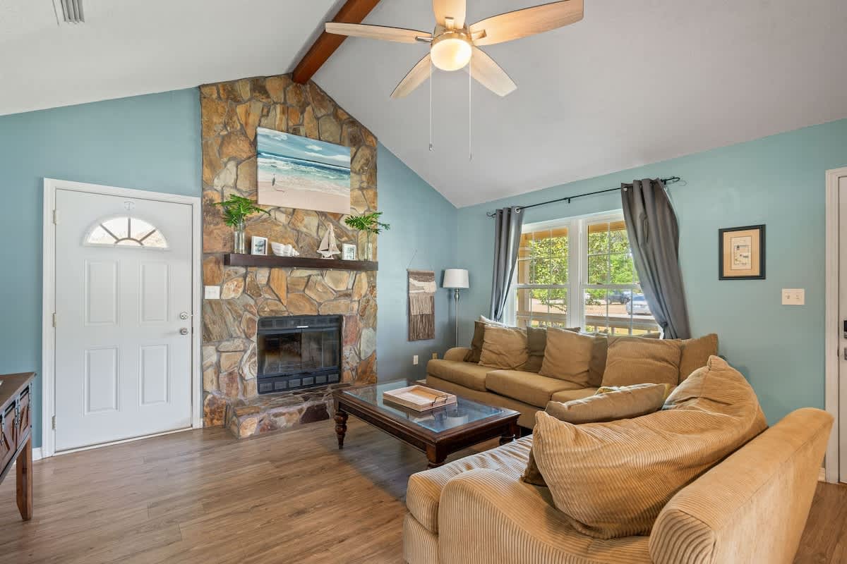 Spacious living room featuring a striking stone fireplace with a wooden mantel, light blue walls, and wood-look flooring, with a white front door and a large window providing natural light.