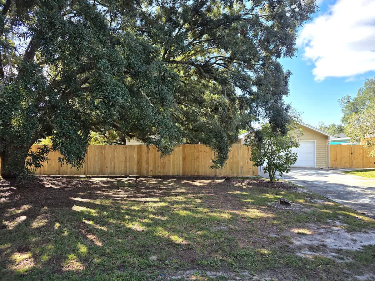 A large, mature oak tree with sprawling branches dominates the foreground, casting shadows on the grassy yard. A newly installed wooden privacy fence runs along the back of the property, with a detached garage visible behind it.