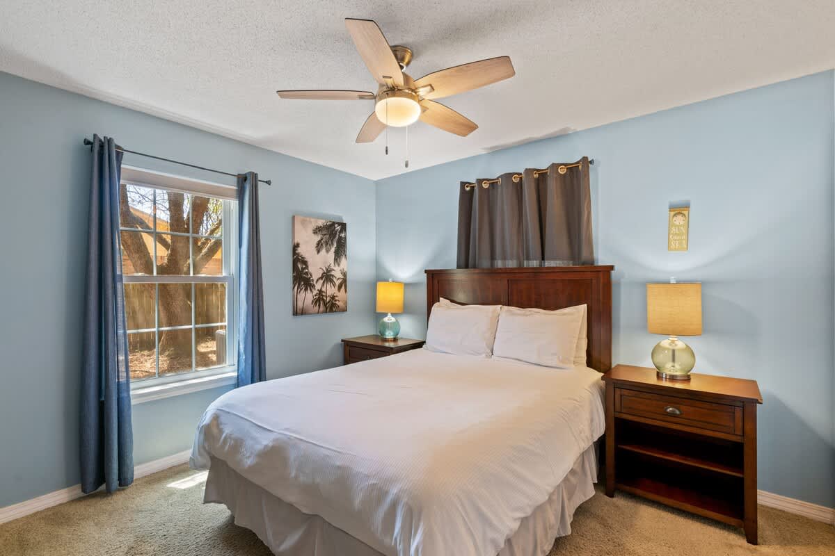 Bright bedroom with light blue walls, a queen-sized bed with white bedding, dark wood nightstands, and a ceiling fan. A large window with blue curtains offers natural light and a view of trees.