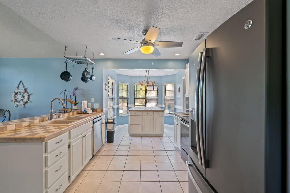 Spacious kitchen with light blue walls, white cabinets, and checkered tile countertops, featuring a central island with a matching tile top and a view into a dining nook with bay windows.