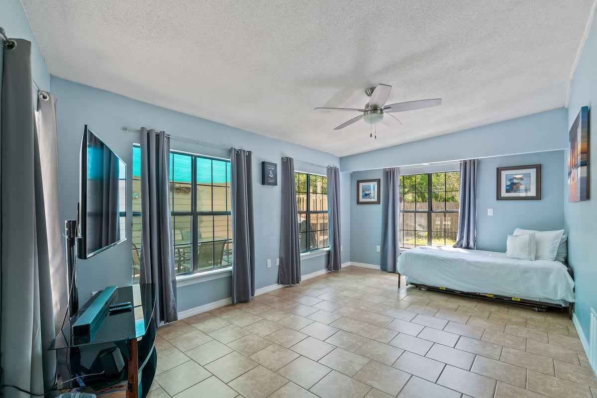 Expansive living room featuring light blue walls, tile flooring, and multiple large windows with gray curtains, furnished with a daybed and a television, illuminated by a central ceiling fan.