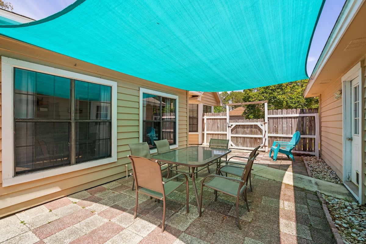 Outdoor patio area with a glass-top dining table and six chairs, covered by a large turquoise shade sail. The space features brick pavers and a wooden fence in the background.