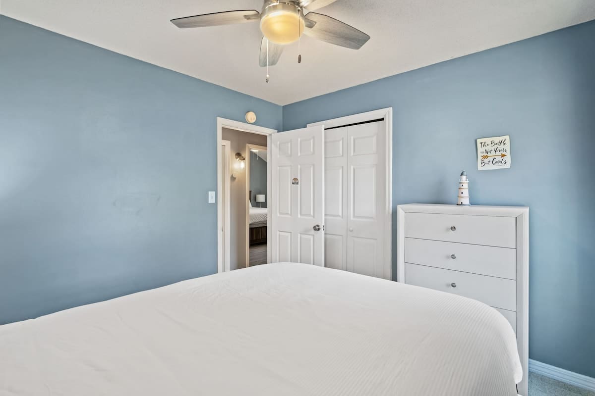 View of bedroom-3 with light blue walls, a white bi-fold closet door, and a white dresser with a lighthouse decoration. A doorway leads to another part of the house.