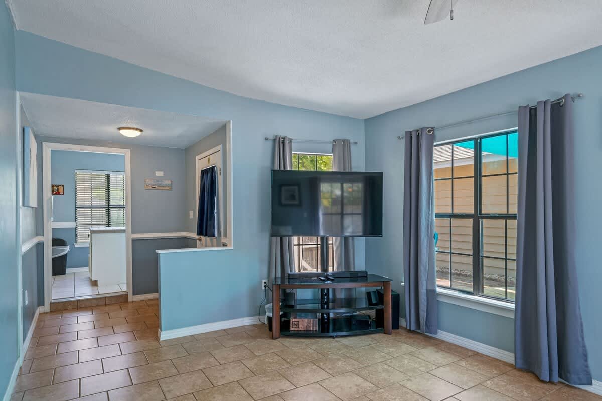 Cozy living area with light blue walls and tile flooring, featuring a large wall-mounted television above a media console, and windows dressed with gray curtains, leading to another room through an open archway.