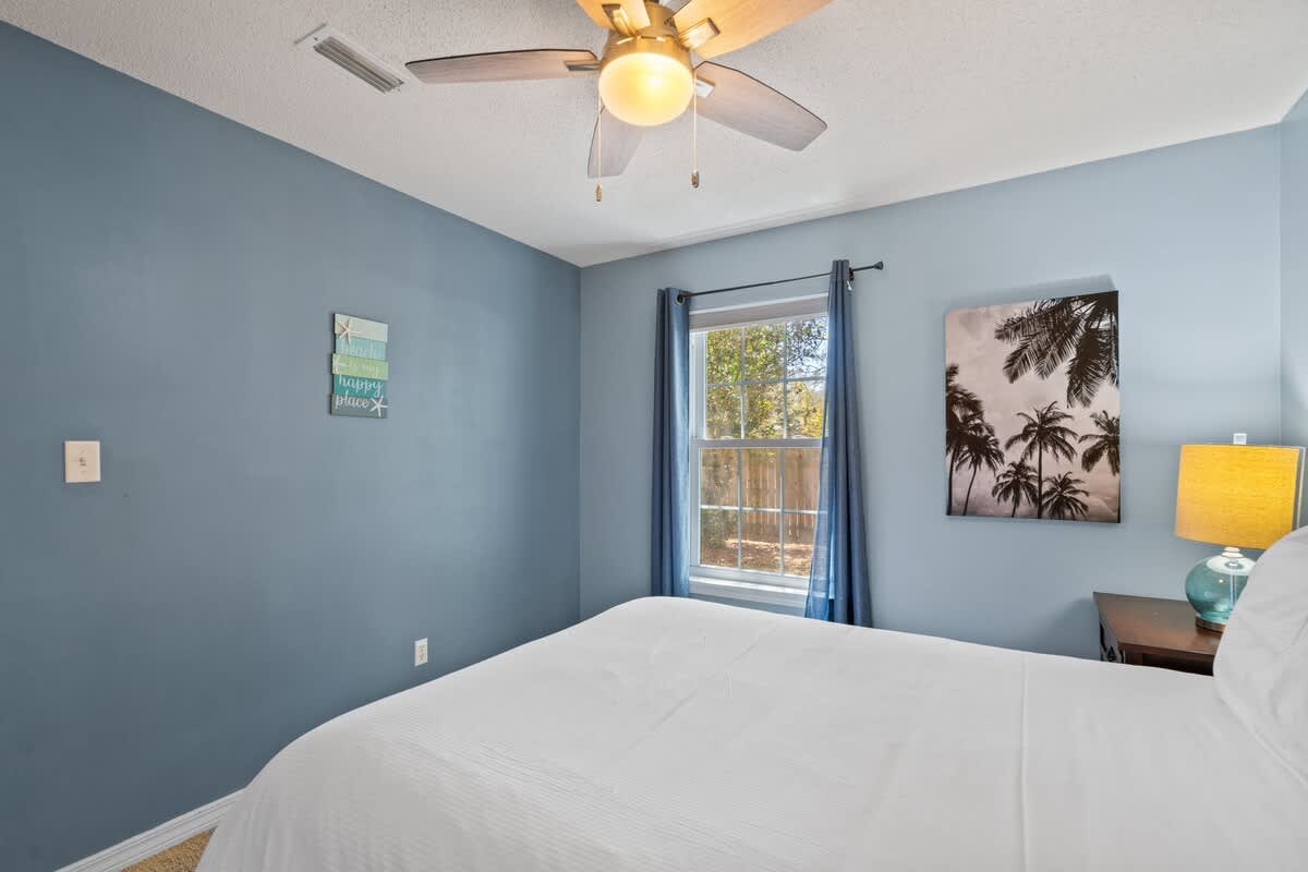 Another view of bedroom-1, featuring light blue walls, a window with blue curtains, and a framed black and white palm tree artwork above a dark wood nightstand with a lamp.