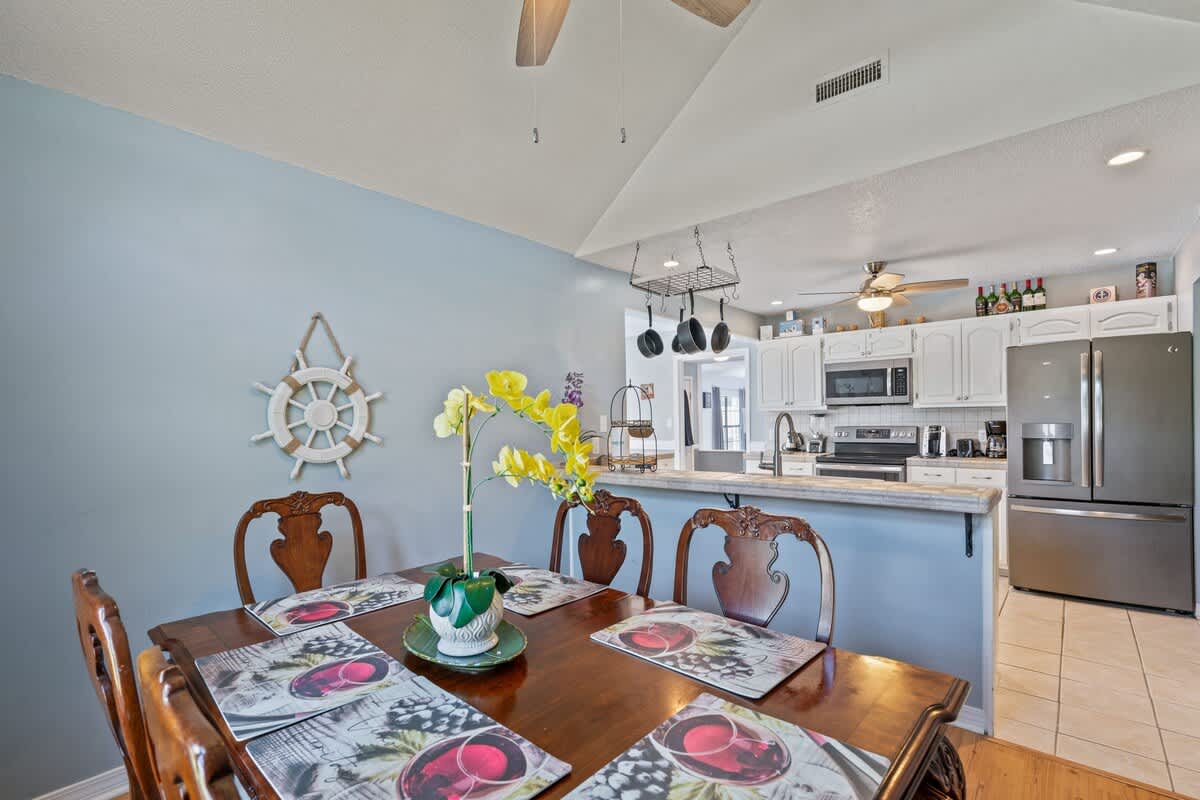 Dining area with light blue walls and hardwood floors, featuring a dark wood dining table, adjacent to a kitchen with white cabinets and stainless steel appliances, separated by a half-wall with a counter.