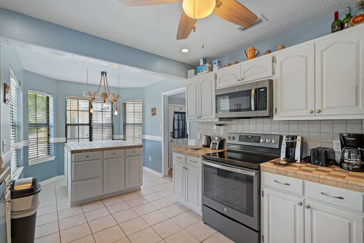 Well-equipped kitchen with white cabinets, tiled countertops, and stainless steel appliances including a microwave and range, featuring a ceiling fan and a tiled floor, with a view into a bright nook.