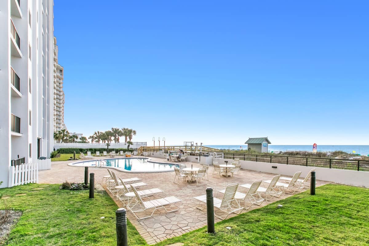 Wide shot of the white high-rise condominium building's exterior, featuring a communal pool area with lounge chairs and tables, a grassy lawn, and the ocean in the background under a bright blue sky.