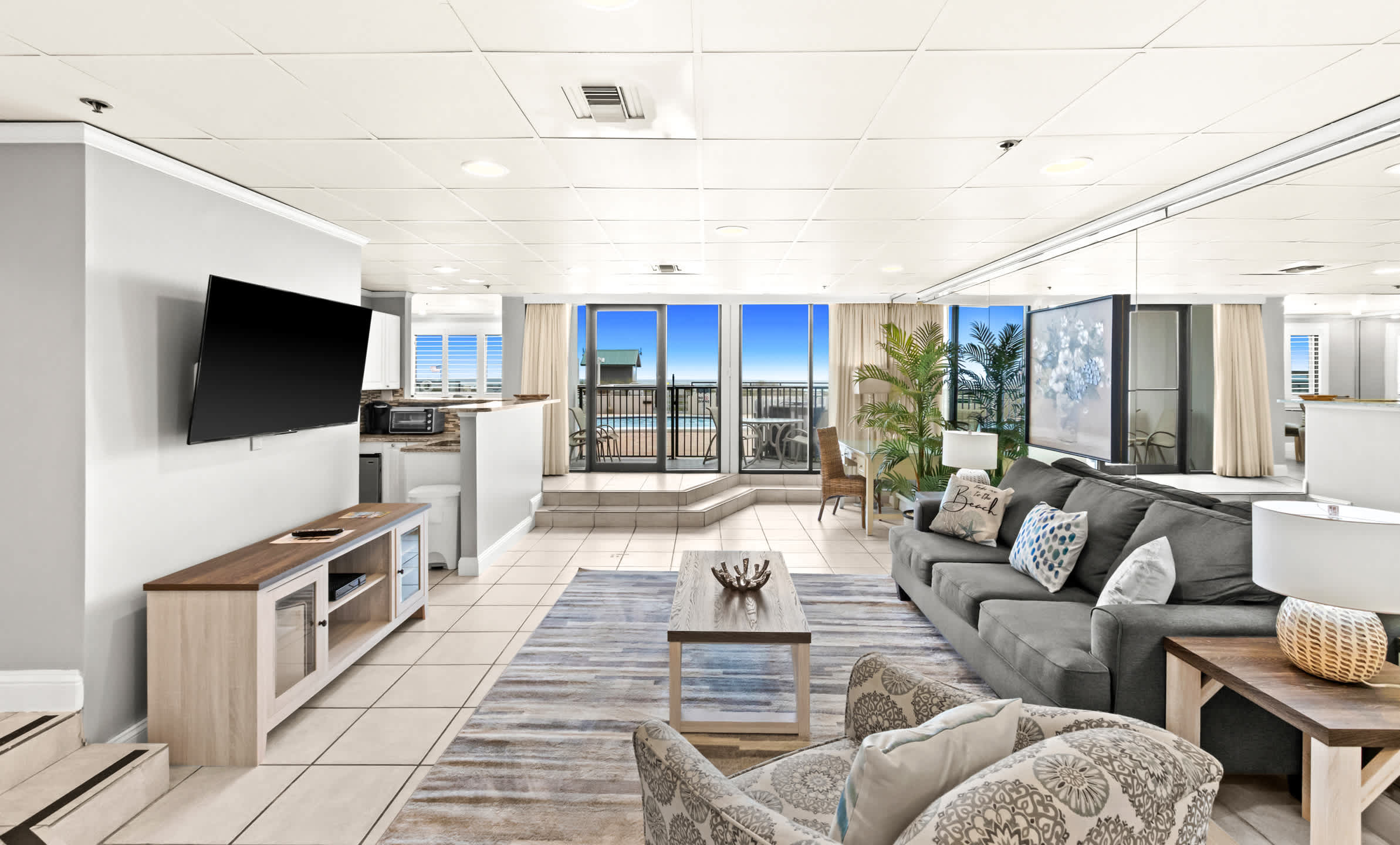 Wide view of the living room with a gray sofa, patterned armchair, and a wooden coffee table, extending towards a kitchen area and a balcony with expansive ocean and pool views.