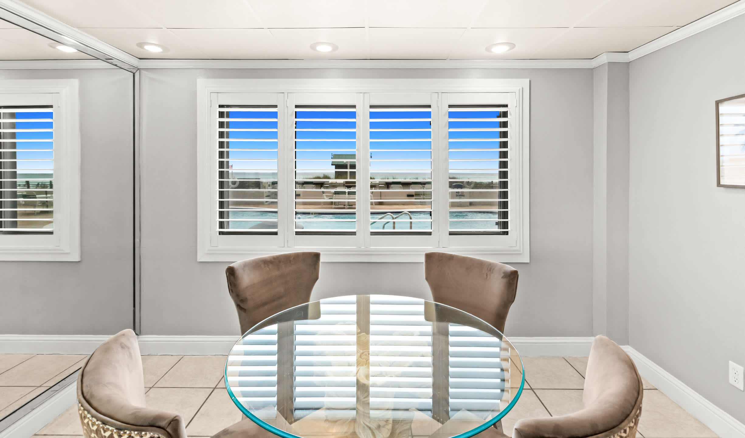 A bright dining area features a round glass-top table with a green base, surrounded by four upholstered chairs, set against a light gray wall with a large window offering a view of a swimming pool and the ocean beyond. Mirrored walls on either side reflect the room, enhancing the sense of space.