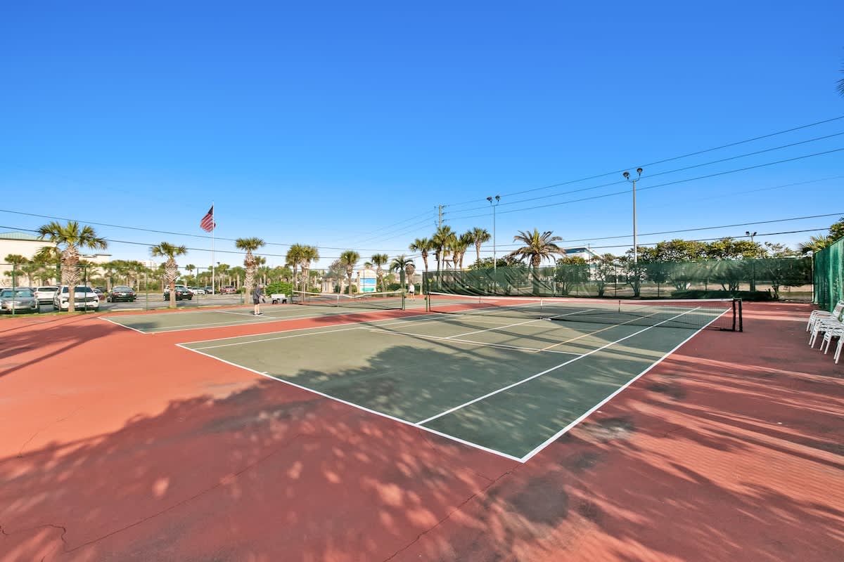 Outdoor tennis court with a green playing surface and red surrounding area, enclosed by a chain-link fence, with palm trees and other buildings visible in the background under a bright blue sky.