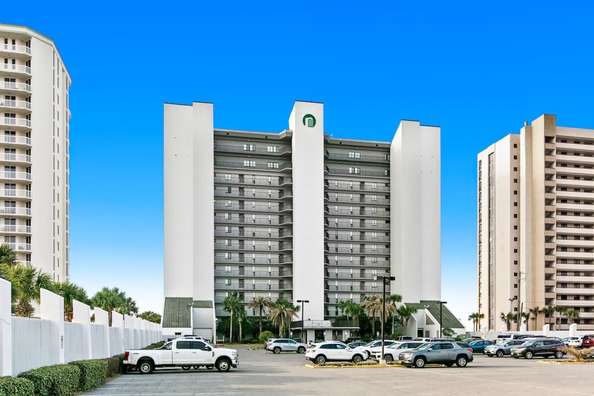 Front view of a tall, modern white condominium building with a green logo at the top, surrounded by a paved parking lot filled with cars and several palm trees under a bright blue sky.