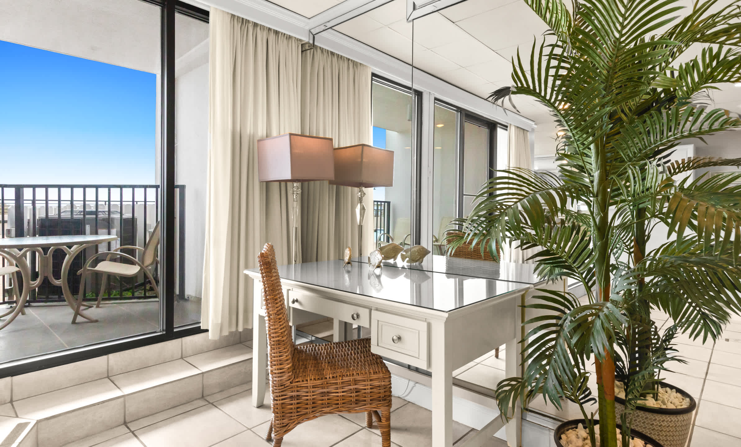 A close-up of the white desk with a wicker chair and a double-shaded lamp, positioned next to sliding glass doors that open to a balcony with outdoor seating and a view of the pool.
