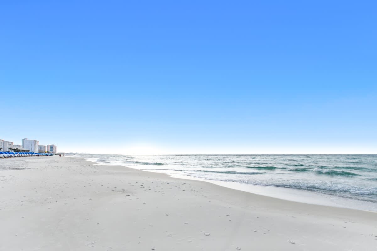 Expansive view of a pristine white sand beach with gentle turquoise waves rolling onto the shore, and distant high-rise buildings visible along the coastline under a bright blue sky.