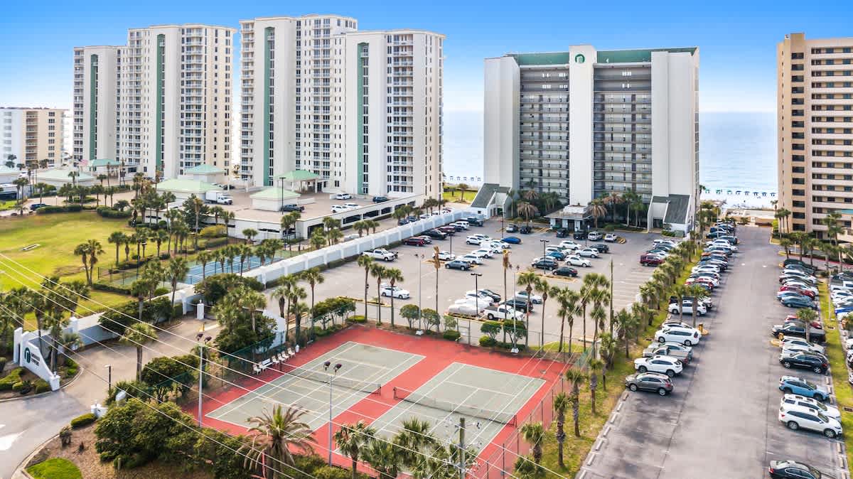 An aerial view showcases a large condominium complex with multiple high-rise buildings, extensive parking lots filled with cars, several red and green tennis courts, and lush green lawns dotted with palm trees, all set against the backdrop of the ocean.