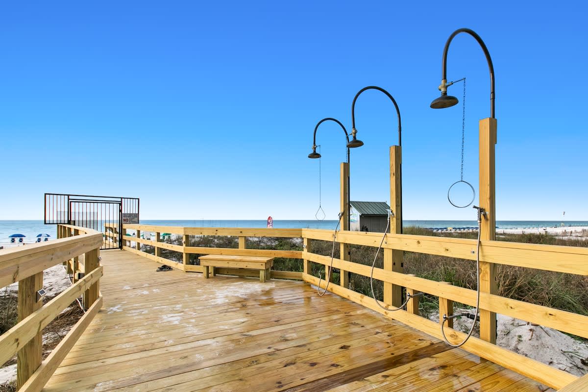 Wooden boardwalk leading towards the beach, equipped with multiple outdoor showers for rinsing off sand, set against a backdrop of the ocean and clear blue sky.