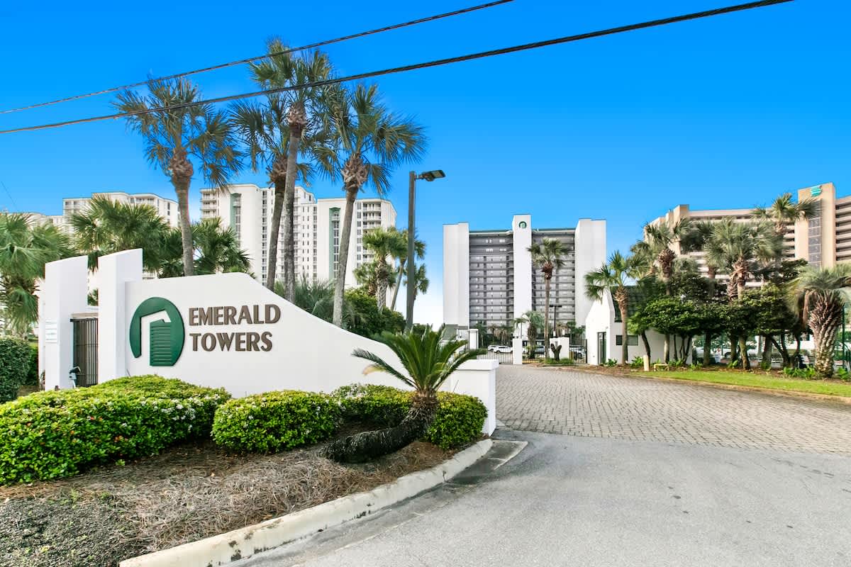 The grand entrance to "Emerald Towers" is marked by a large white sign with green lettering, surrounded by lush green landscaping and palm trees. The multi-story condominium buildings are visible in the background under a clear blue sky.