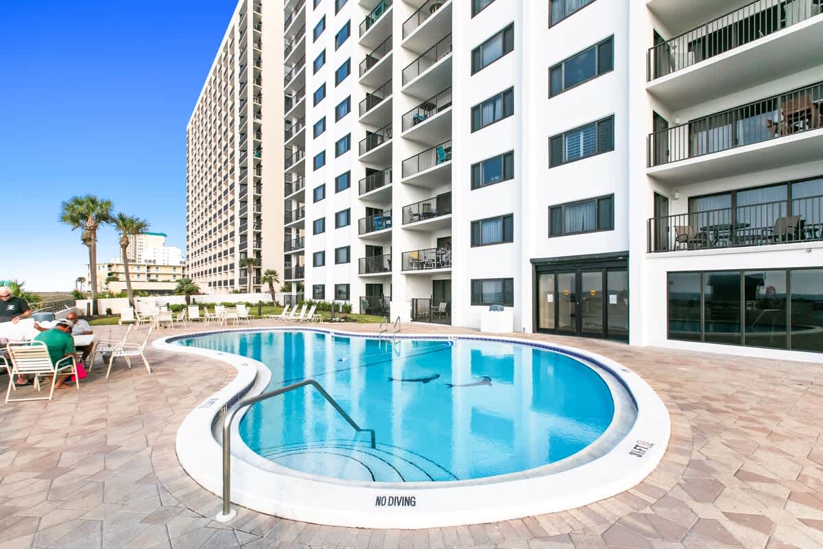 Curved outdoor swimming pool with steps and a handrail, surrounded by a paved deck with lounge chairs, adjacent to a tall white condominium building under a clear blue sky.