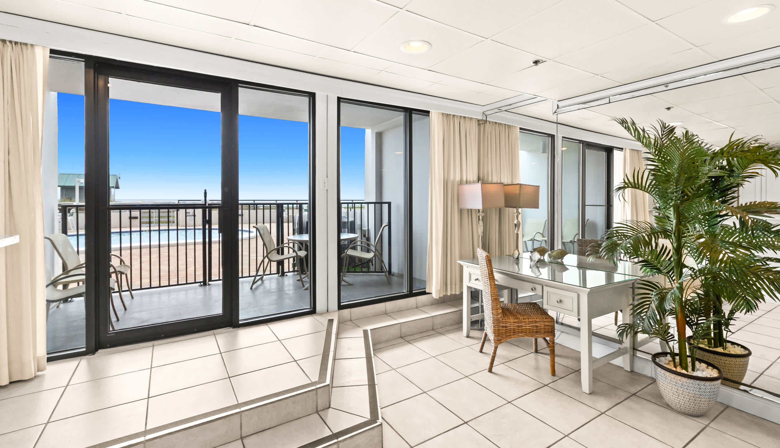A bright corner of the living area with a white desk and wicker chair, illuminated by natural light from sliding glass doors leading to a balcony with views of a pool and the ocean.