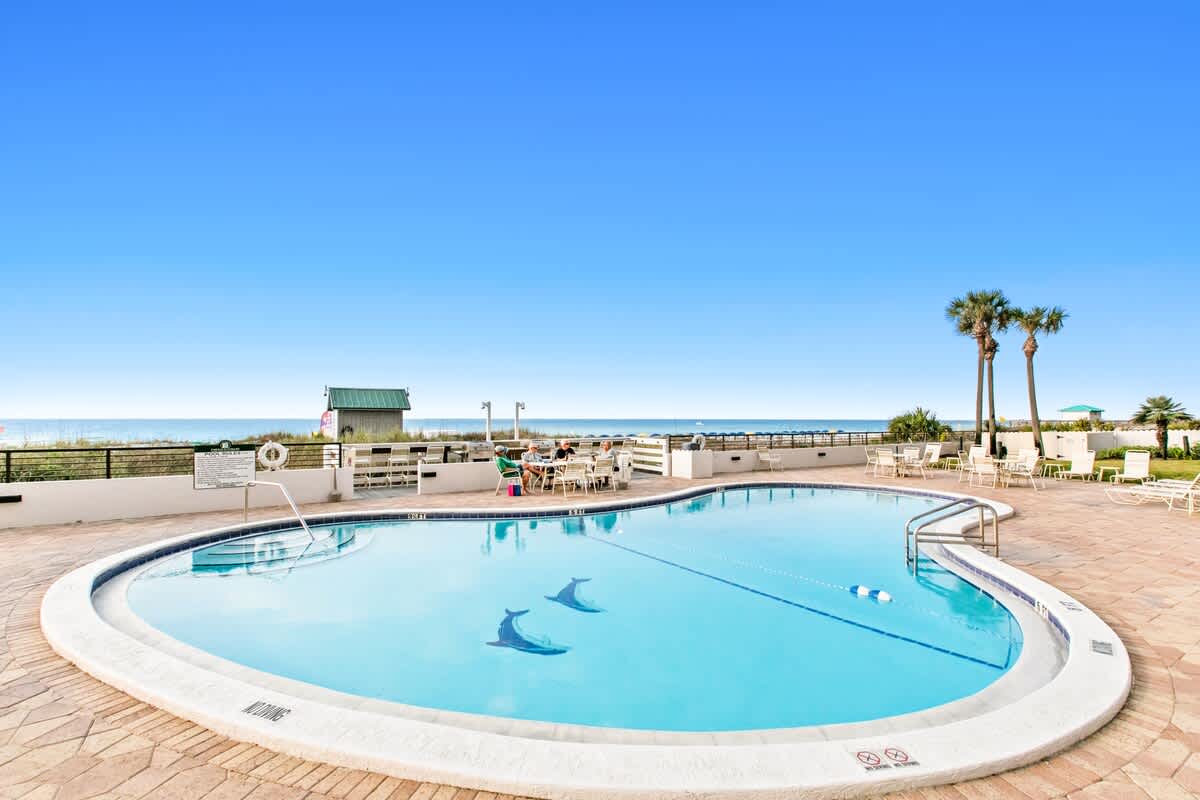 Large, kidney-shaped communal swimming pool with dolphin mosaics on the bottom, surrounded by light brown paver decking and overlooking the ocean under a clear blue sky. Several lounge chairs and dining tables are visible around the pool area.