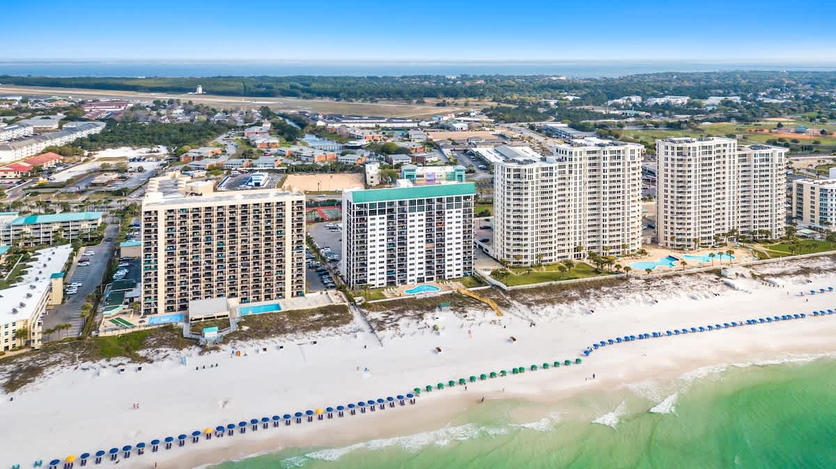 An expansive aerial shot capturing a series of tall beachfront condominium buildings, their associated outdoor pools, and the pristine white sand beach extending along the turquoise coastline. The surrounding urban landscape is also visible.