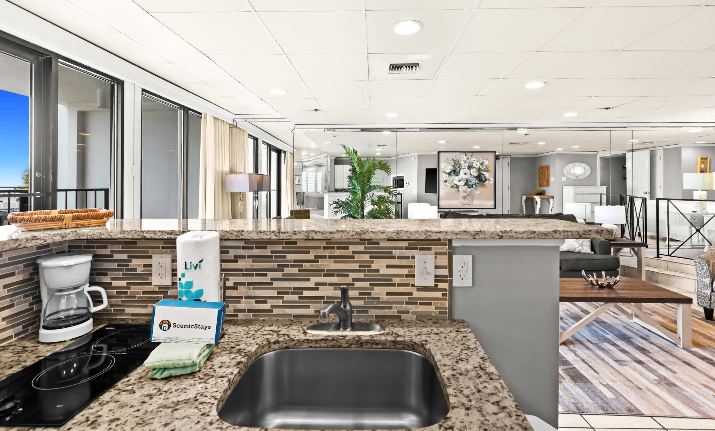 A wider view of the kitchenette counter with a small sink, stovetop, and coffee maker, featuring granite countertops and a mosaic tile backsplash, overlooking the living area and a mirrored wall.