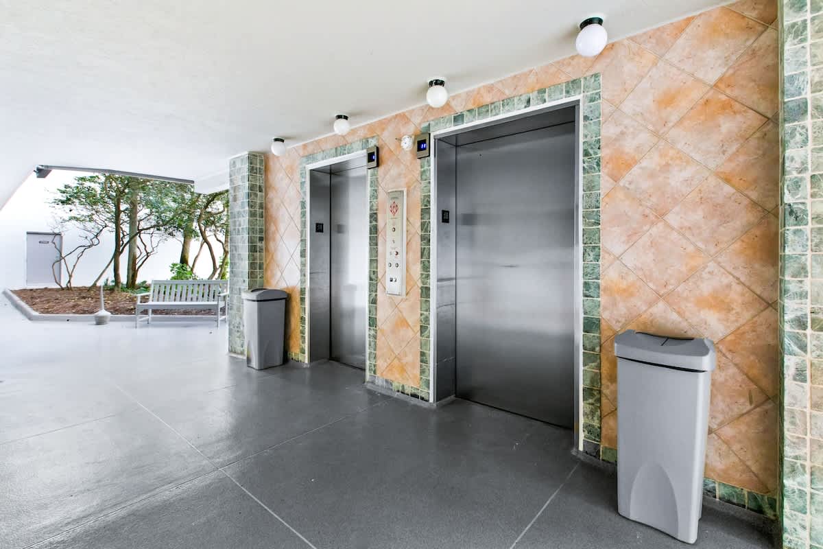 Interior lobby area featuring two stainless steel elevators, walls adorned with patterned green and orange tiles, and a light gray tiled floor, with a trash can beside each elevator.