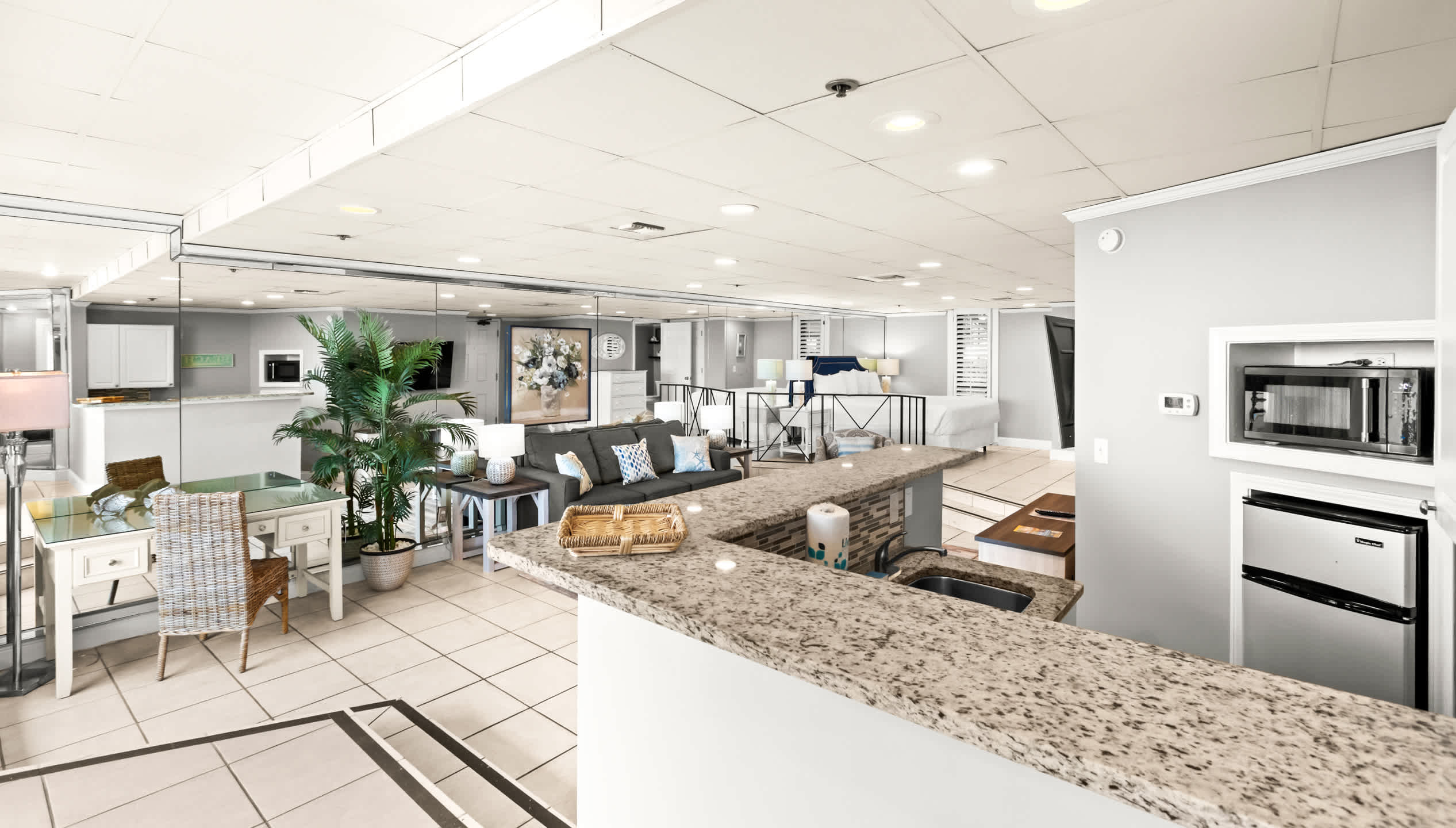 An expansive view of the living area from the kitchenette, highlighting the open layout with a gray sofa, armchair, and a desk area, all reflected in a large mirrored wall.