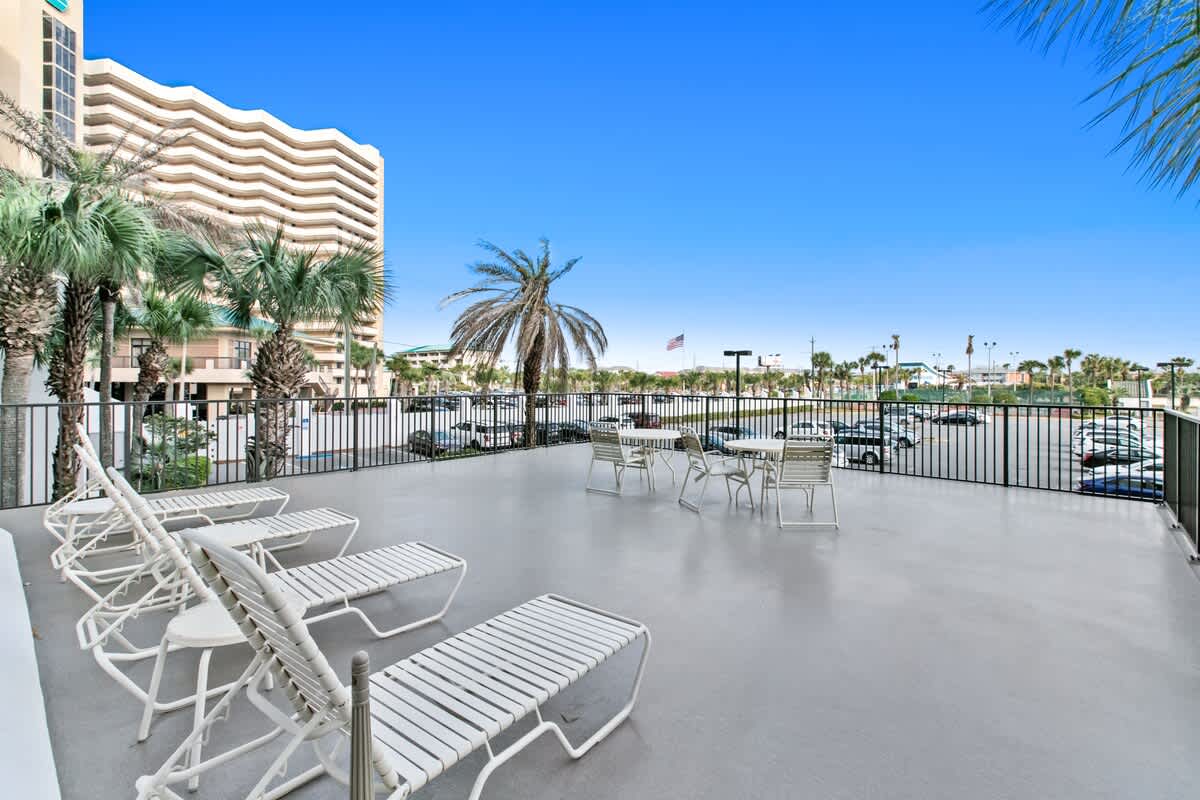 An outdoor communal deck furnished with white lounge chairs and dining tables, offering views of a parking lot, palm trees, and adjacent buildings under a clear blue sky. A black metal railing encloses the space.