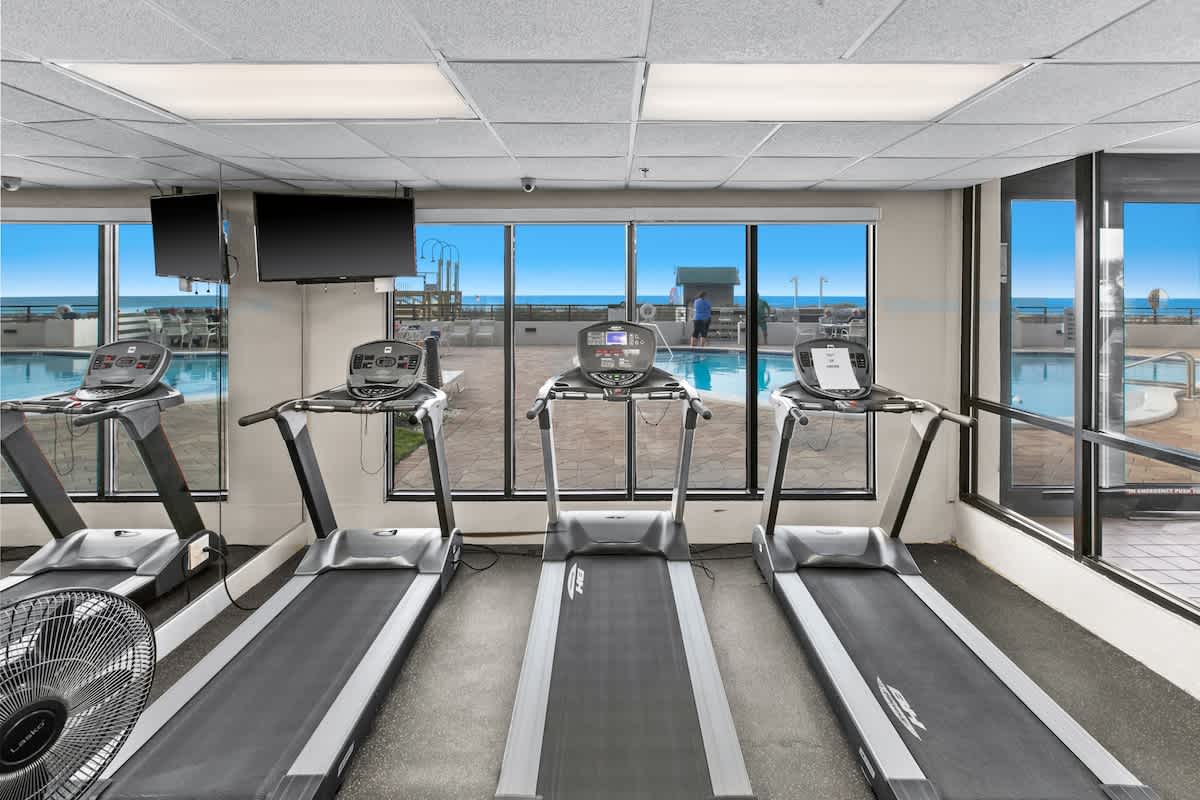 Three treadmills are lined up facing expansive windows, offering a direct view of an outdoor swimming pool and the vast ocean beyond. A ceiling-mounted TV is visible above the windows.
