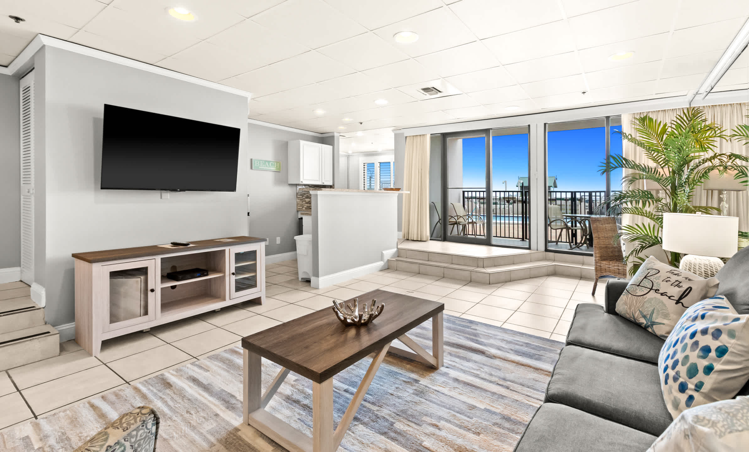 Living room with a large wall-mounted TV above a light wood media console, a gray sofa with decorative pillows, and a glimpse of the balcony with outdoor seating.
