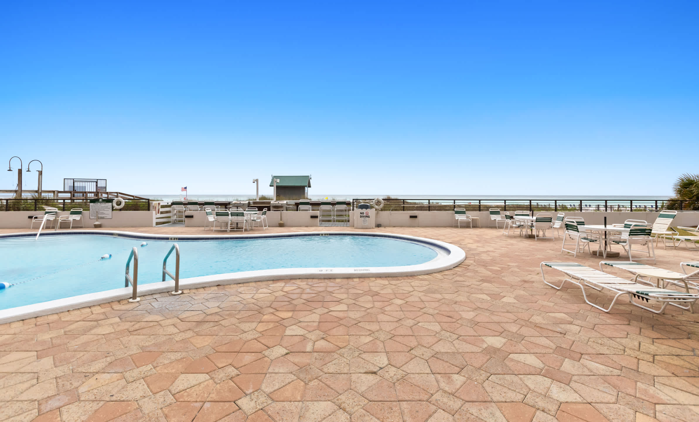 This view captures the communal swimming pool and its surrounding stone patio, furnished with numerous lounge chairs and dining sets, all facing the expansive ocean horizon. A small building and palm trees are visible near the beach access.
