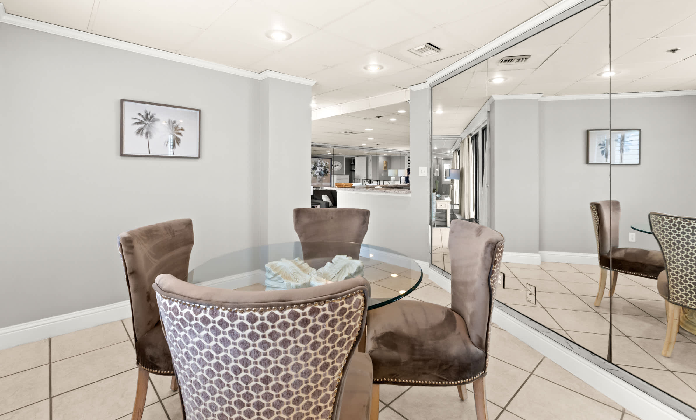 This dining area showcases a round glass-top table with a decorative centerpiece, flanked by four plush upholstered chairs, two with patterned backs, all on a tiled floor. Mirrored walls expand the view into an adjacent living space, creating an open and airy feel.