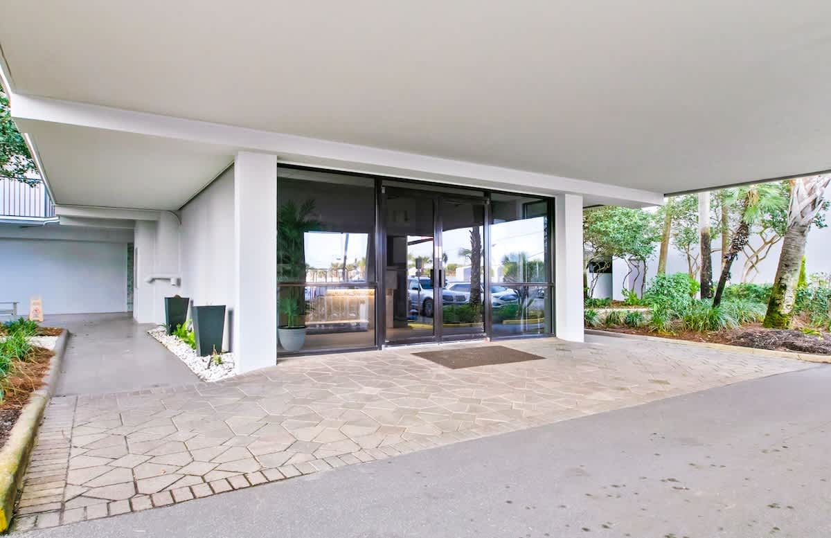 Covered entrance area of a building with large glass double doors, a paved walkway, and lush green landscaping with palm trees, leading into the main lobby.