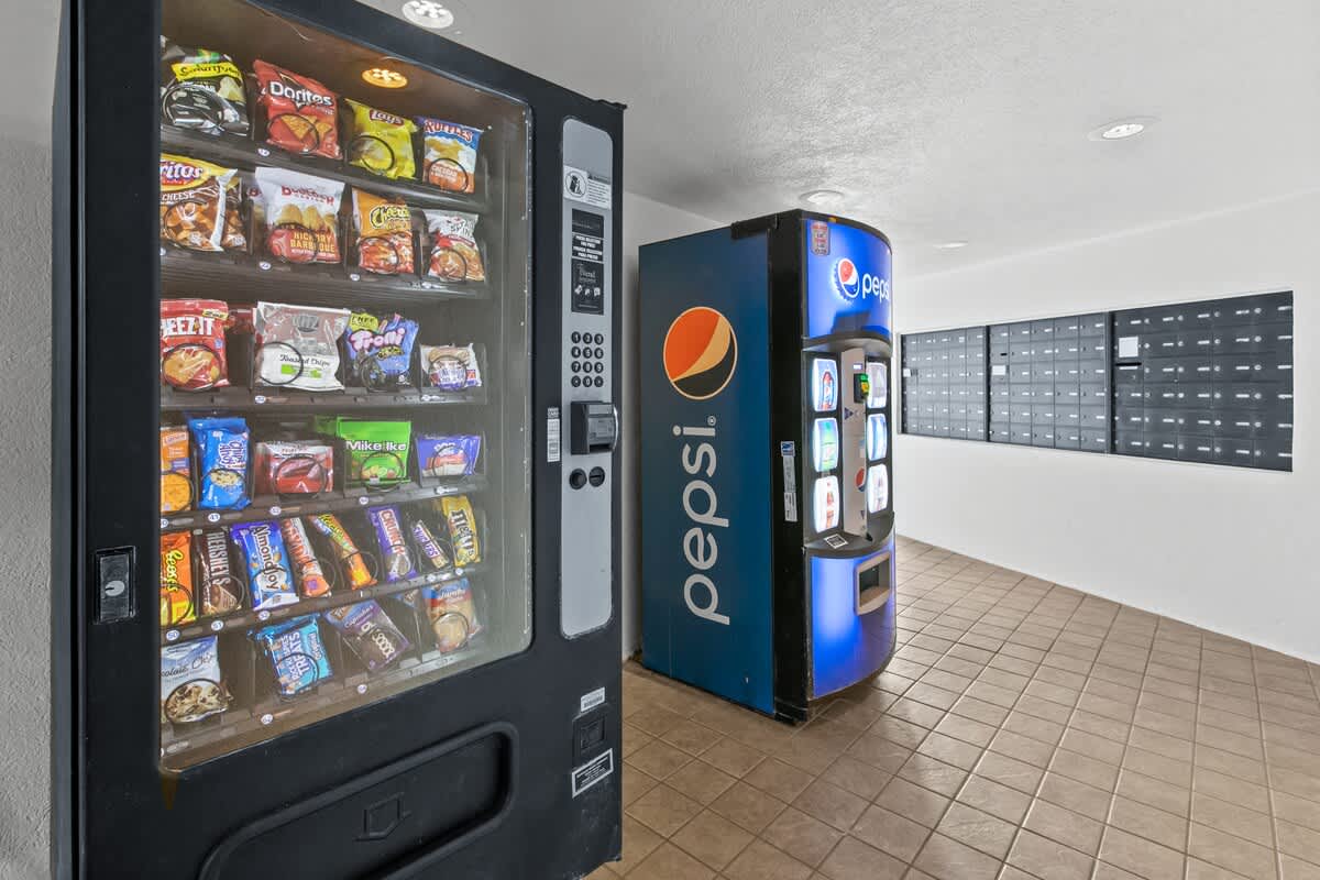 Common area with a snack vending machine and a Pepsi drink vending machine, alongside a wall filled with mailboxes, all under recessed lighting.