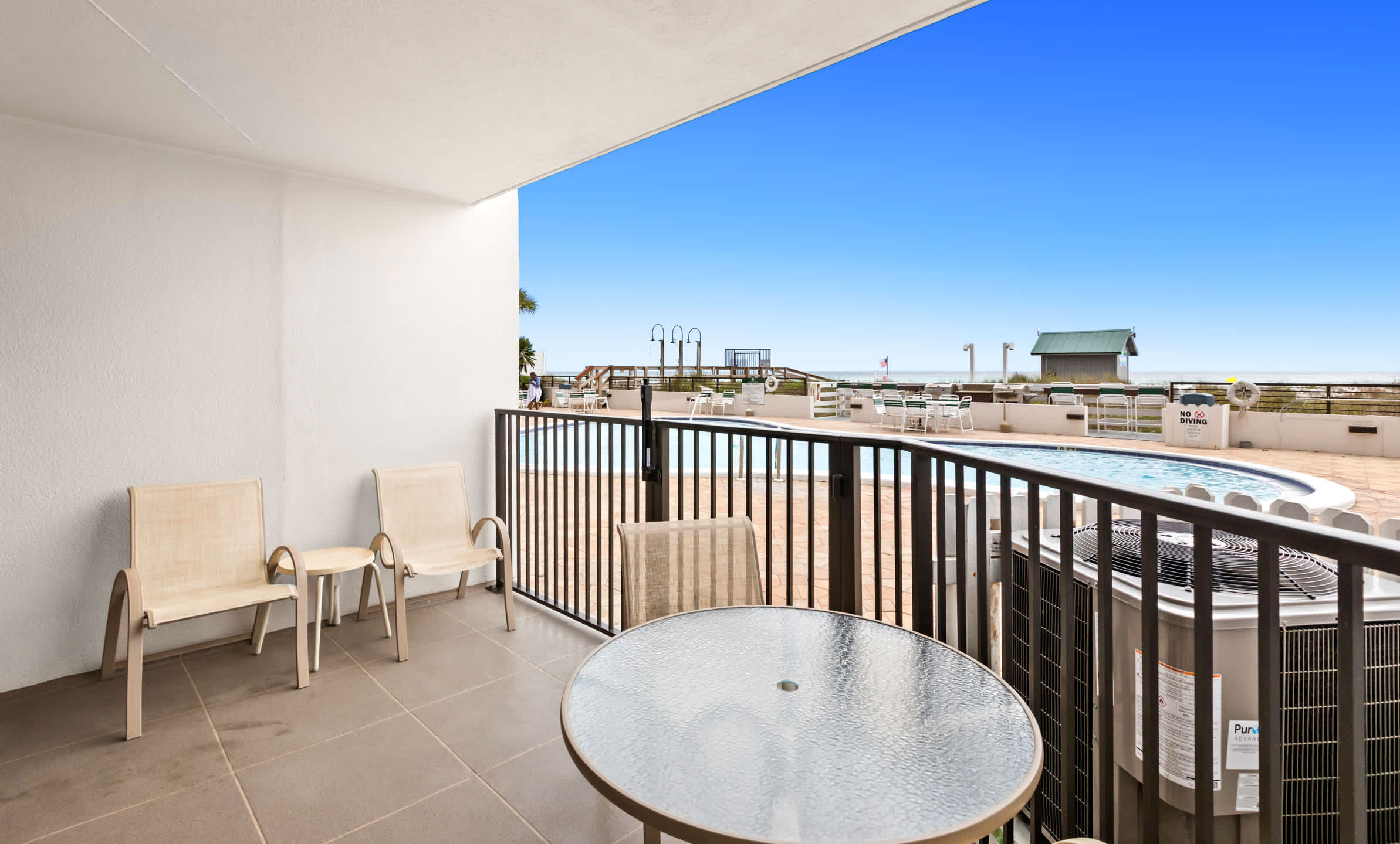 This spacious balcony features a small round glass-top table with two chairs, plus two additional lounge chairs, offering a direct view of the communal pool and the ocean beyond. An outdoor air conditioning unit is visible in the corner.