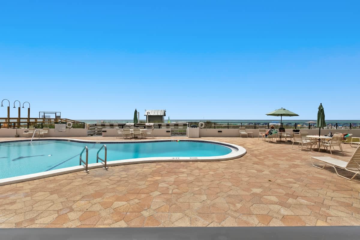 An elevated view of the communal swimming pool and its surrounding patio, filled with lounge chairs and dining tables, all set against the backdrop of the ocean under a bright blue sky. Green umbrellas provide shade for some seating areas.