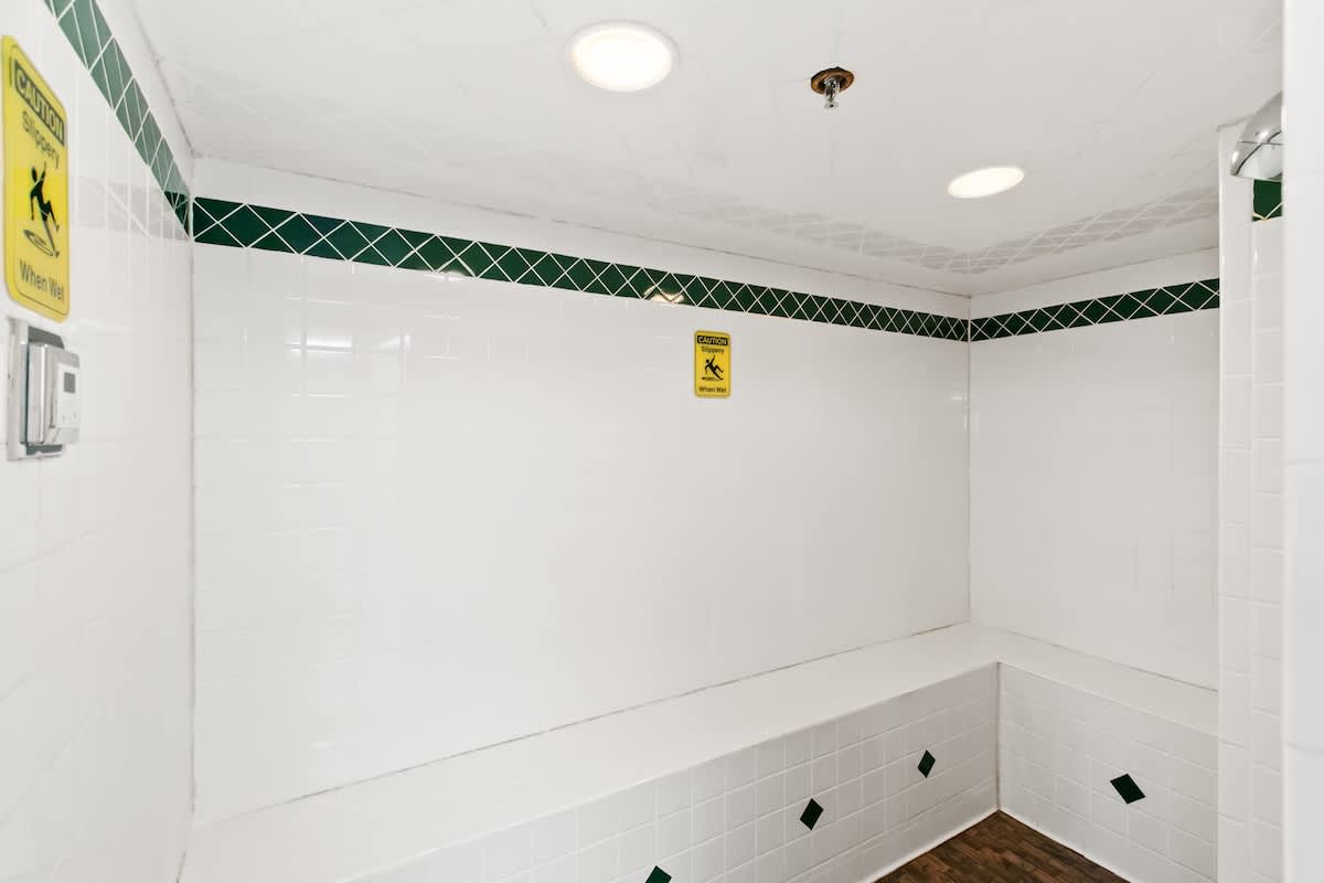 A clean, white-tiled changing room or shower area featuring a built-in bench along the wall and a dark green decorative tile border. "Caution Slippery When Wet" signs are visible.