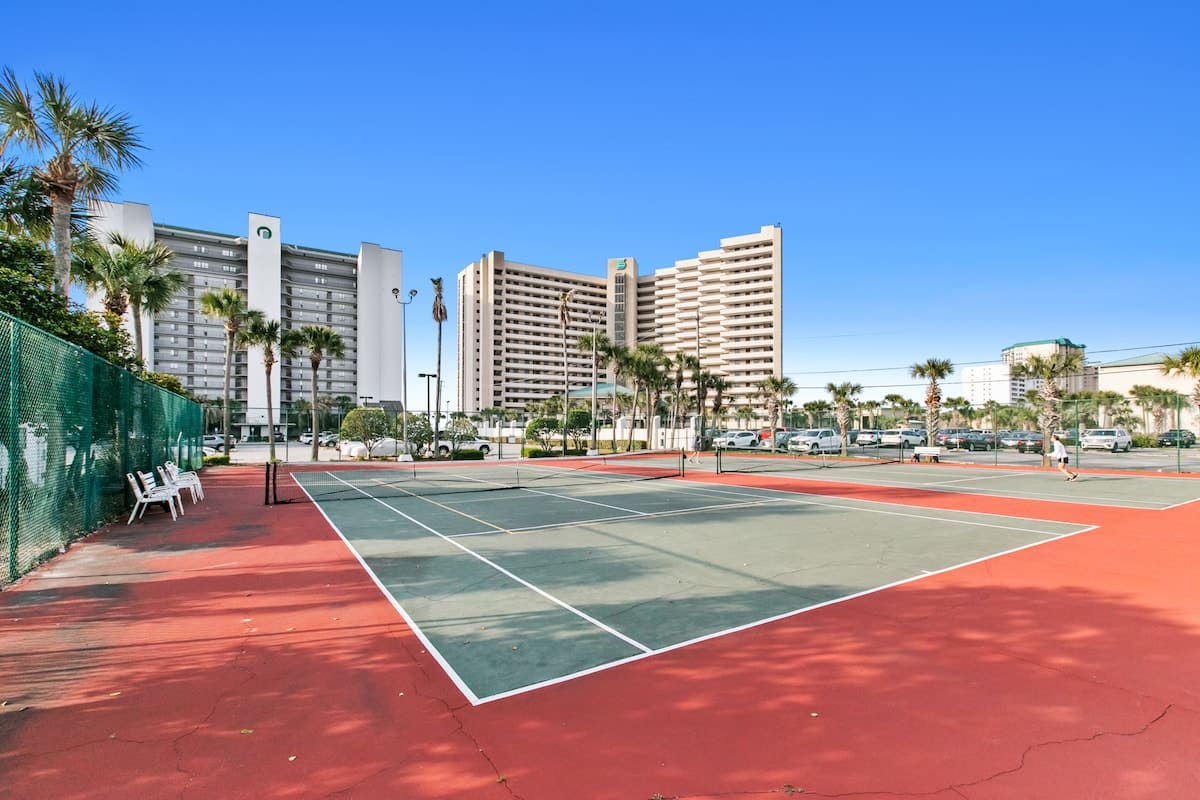 Wide view of two outdoor tennis courts with green and red surfaces, surrounded by a fence, with multiple tall condominium buildings and palm trees in the background under a clear blue sky.