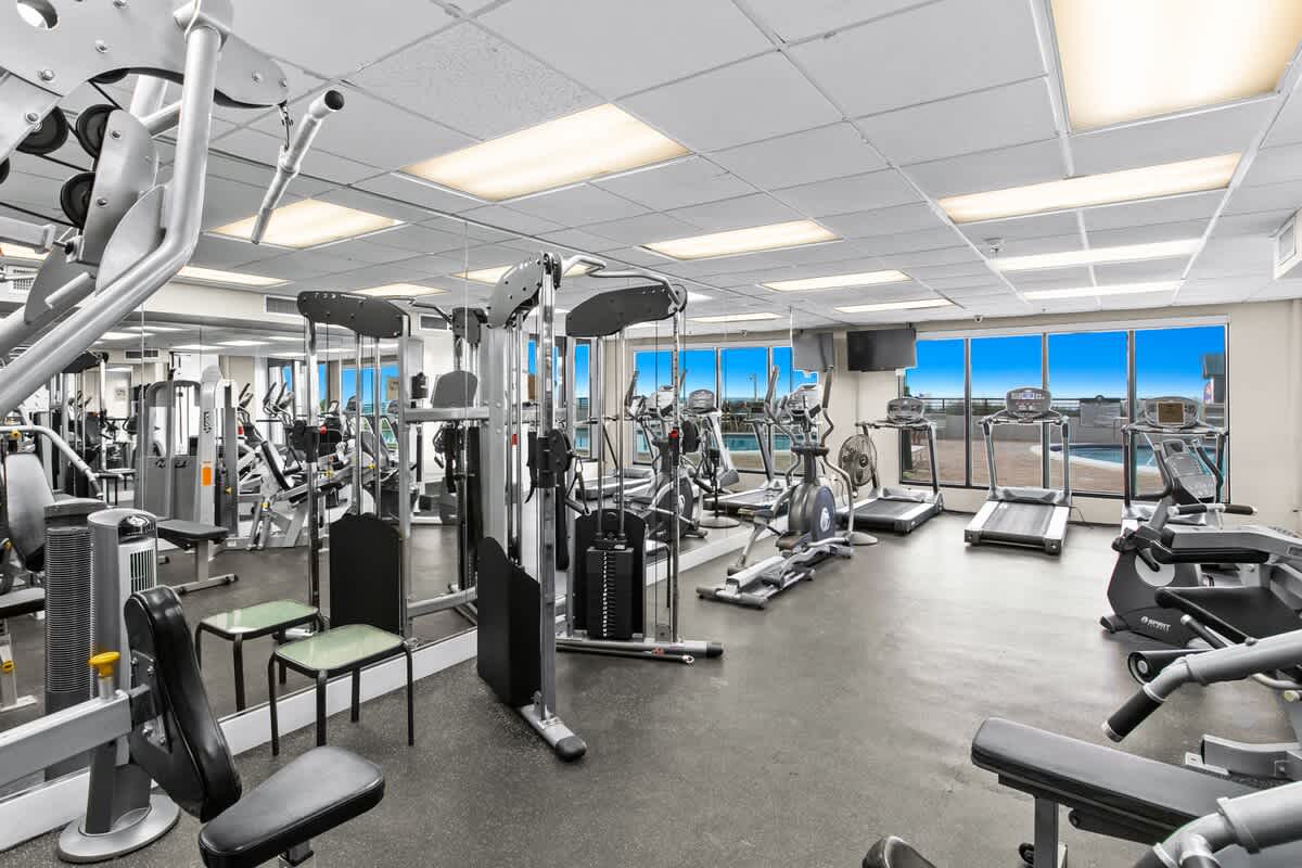 A well-equipped fitness center featuring a variety of cardio and weight machines, with large windows offering views of an outdoor pool and the ocean. The space has a dropped ceiling with fluorescent lighting and mirrored walls.