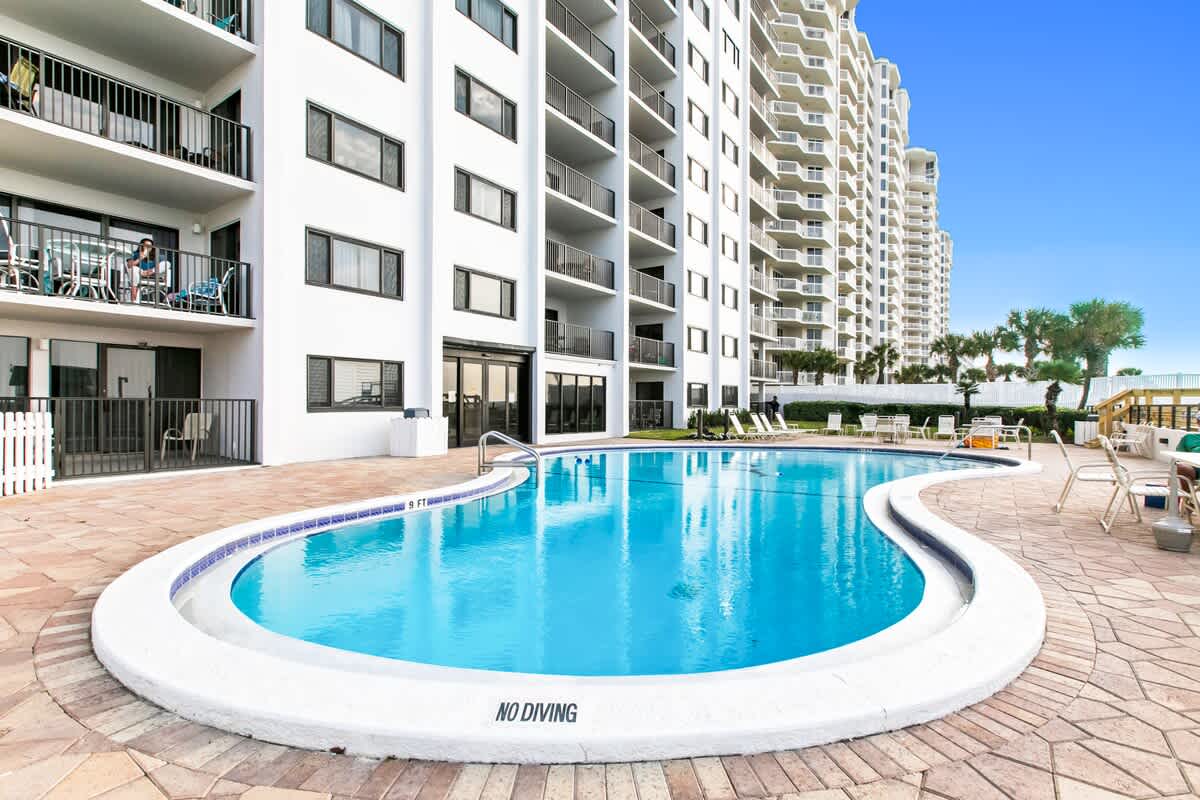 A smaller, kidney-shaped communal swimming pool with a "NO DIVING" sign, surrounded by light brown paver decking, located next to the white condominium building with balconies.