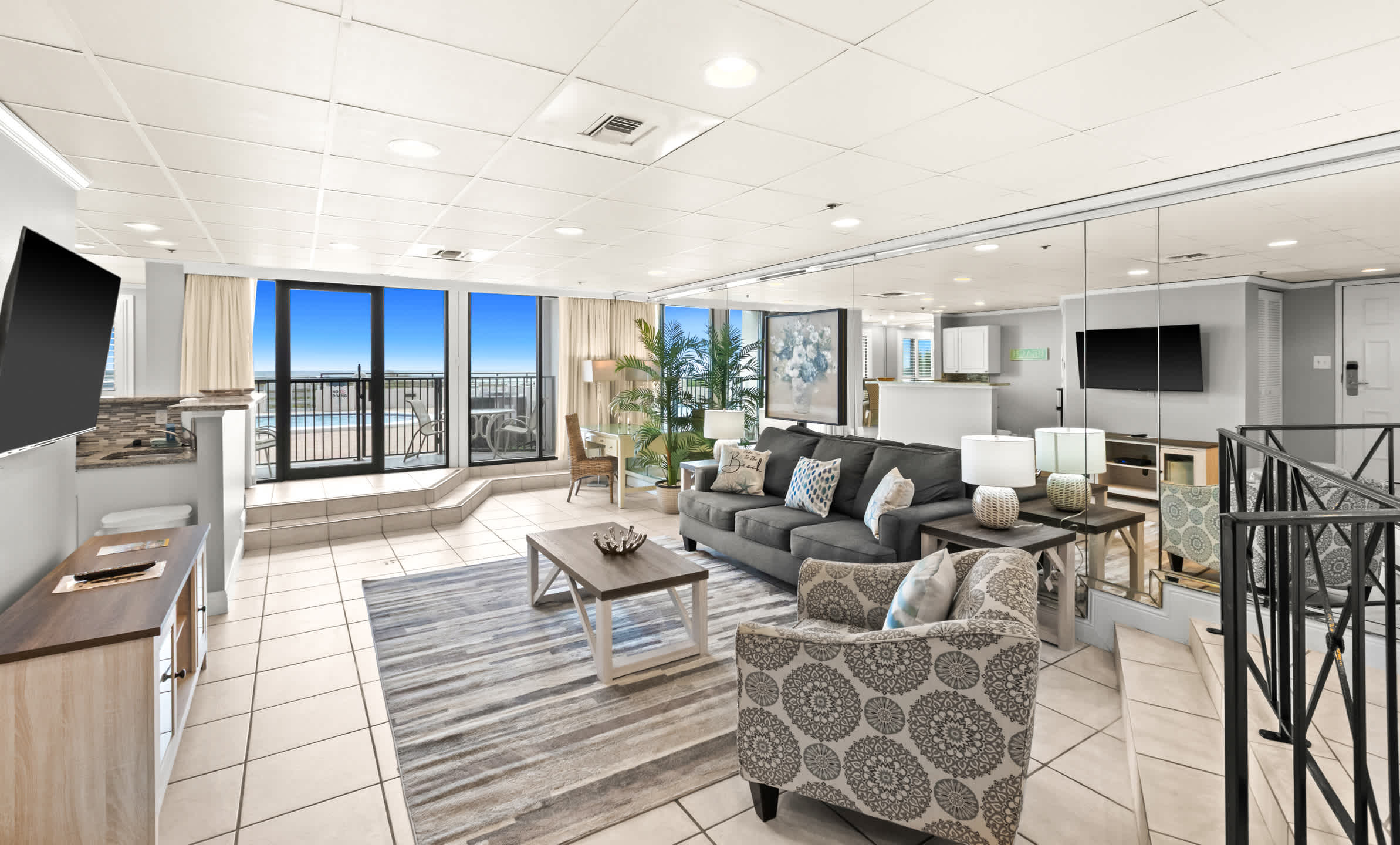 Spacious living room with a gray sofa and patterned armchair, a wooden coffee table on a striped rug, leading to a balcony with a view of the communal pool and ocean.