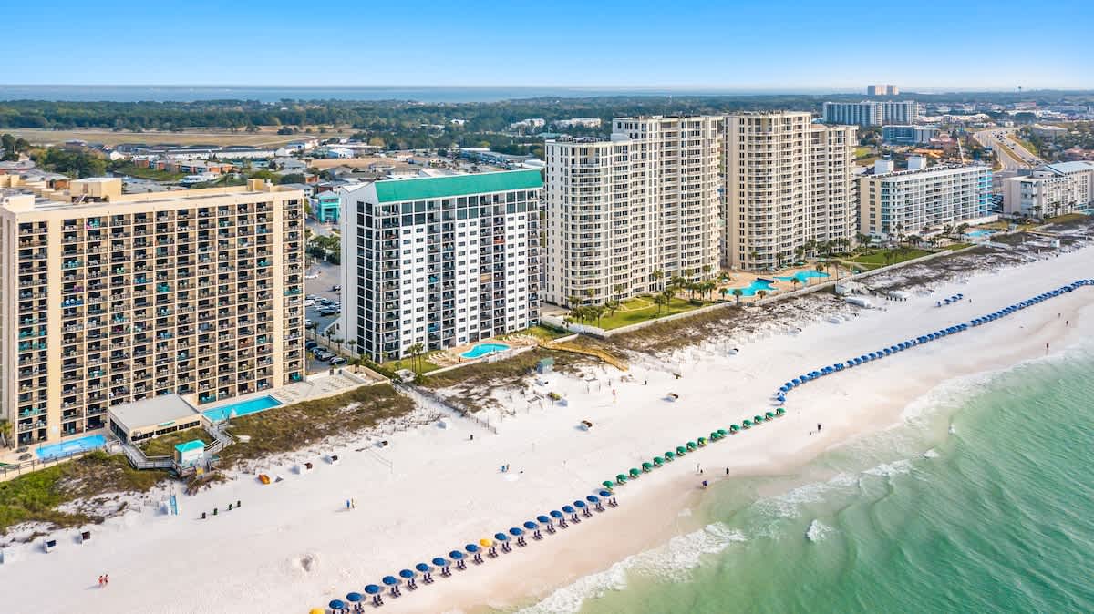 A sweeping aerial view of a vibrant beachfront property featuring multiple high-rise condominium buildings, several outdoor pools, and a long stretch of white sand beach dotted with colorful umbrellas and lounge chairs. The turquoise ocean meets the shore.