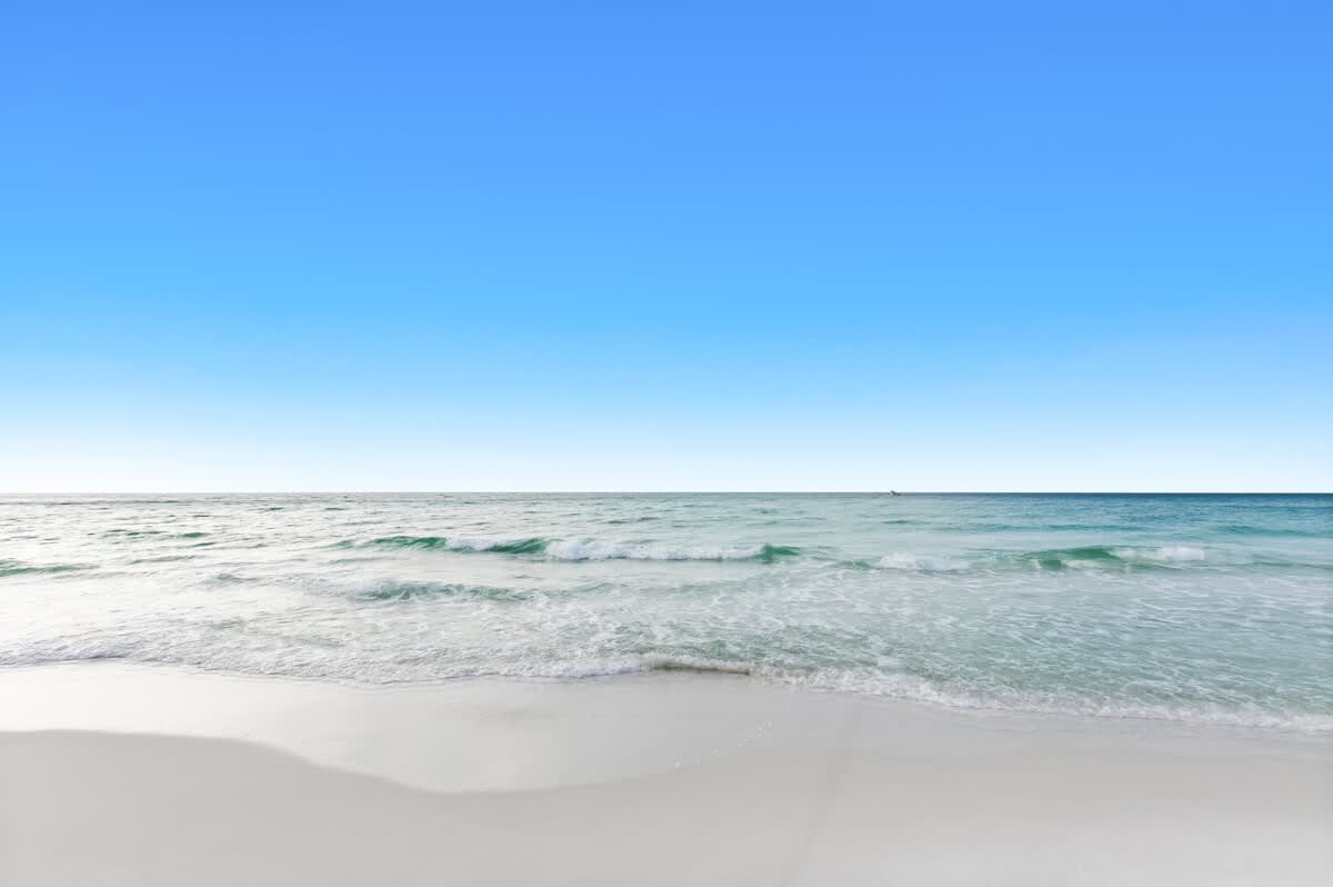 Close-up view of the clear turquoise ocean with small waves breaking on the pristine white sand beach, under a vast, cloudless blue sky.
