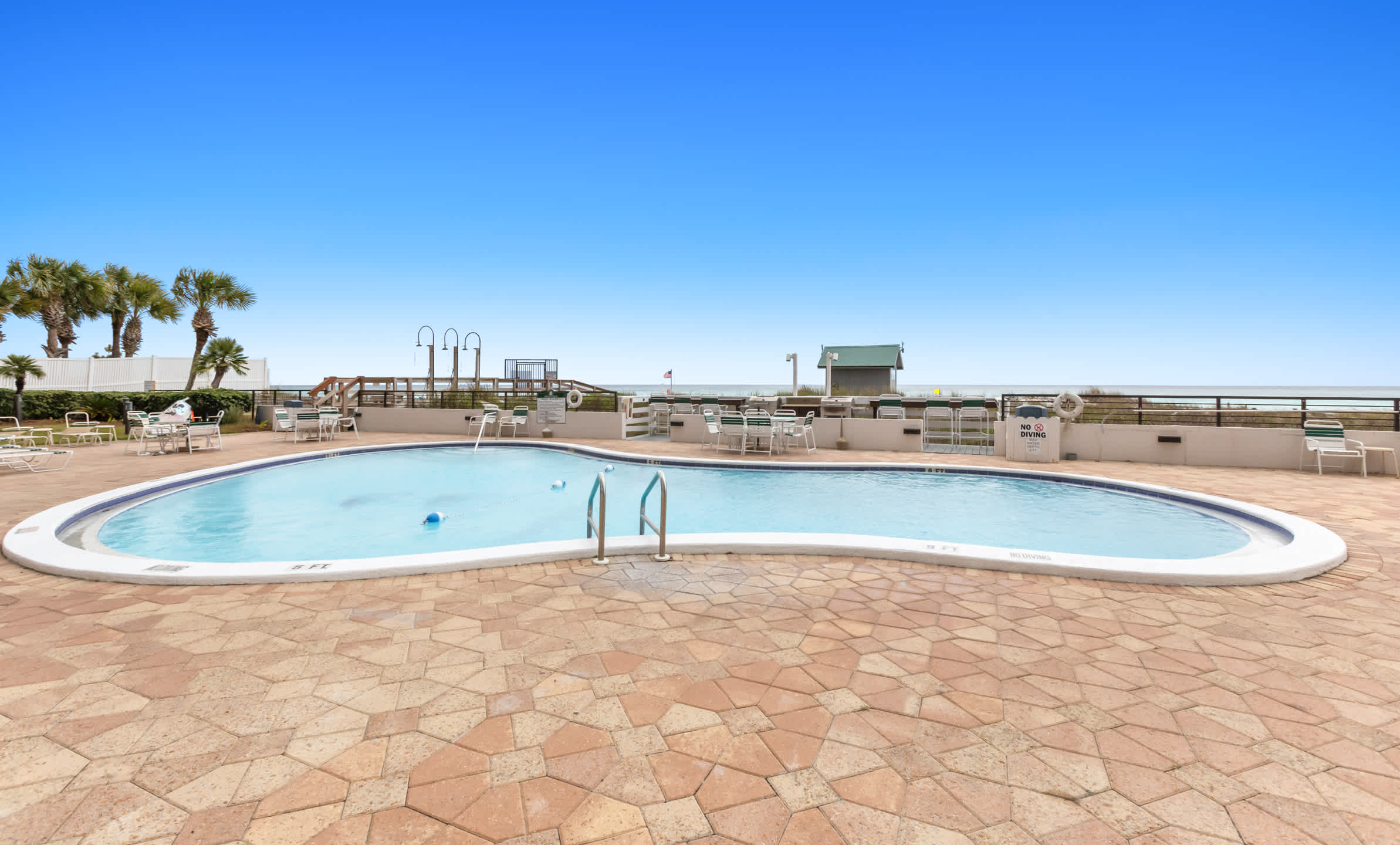 A wide shot of the communal swimming pool shows its irregular shape and blue water, set against a textured stone patio with various lounge chairs and tables. The ocean is clearly visible in the distance, framed by a clear blue sky.