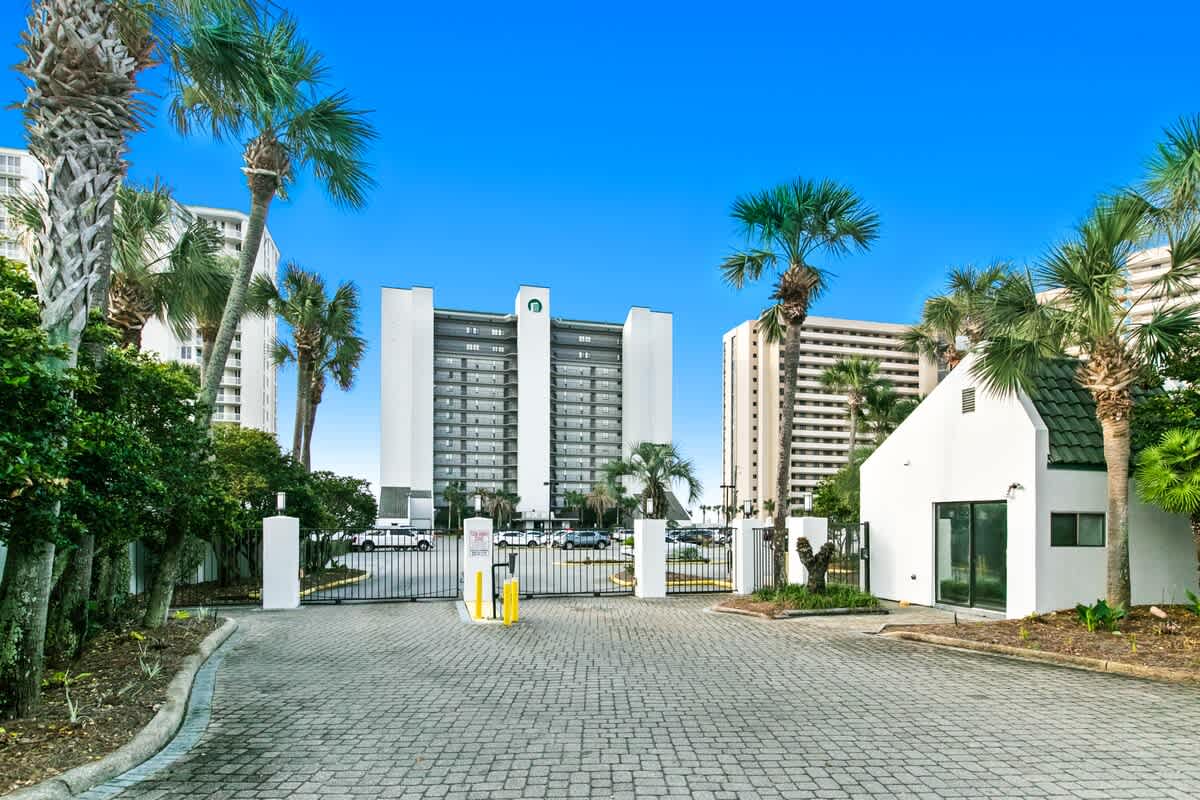 Gated entrance to a condominium complex, featuring a paved driveway, white gate pillars, a small guardhouse, and tall white buildings surrounded by palm trees and lush greenery under a clear blue sky.