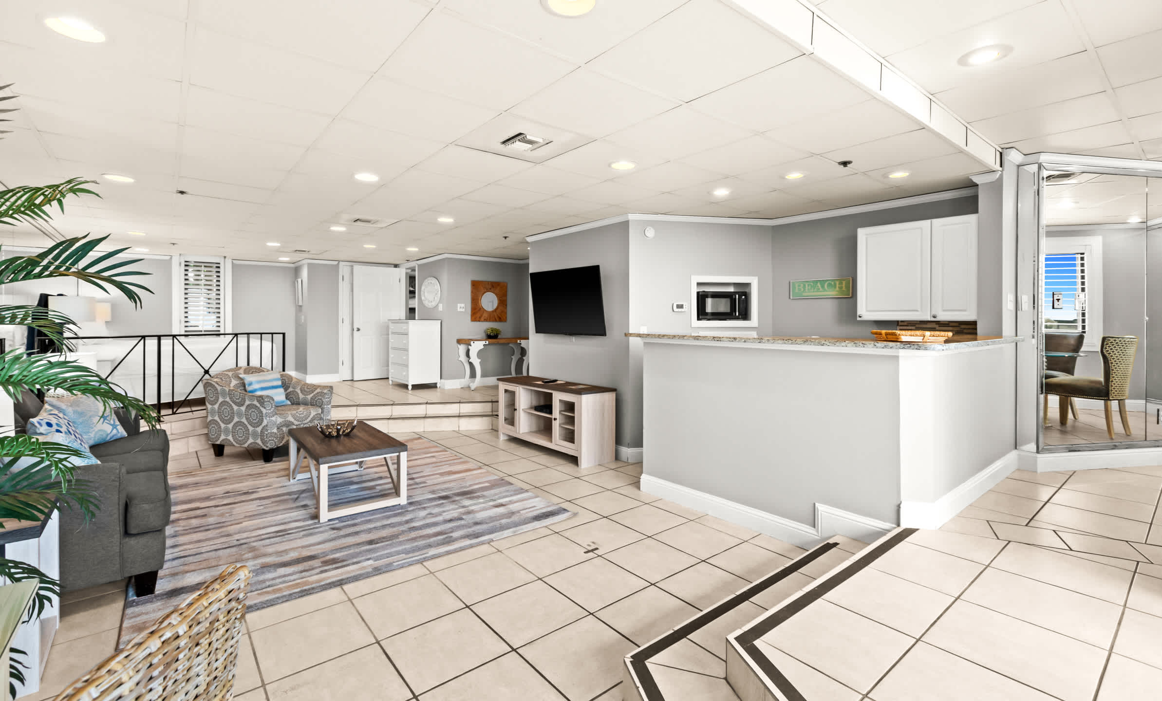 A panoramic view of the open-concept living space, showing a gray sofa, patterned armchair, coffee table, and a TV console, with a kitchenette bar and a dining area visible in the background.