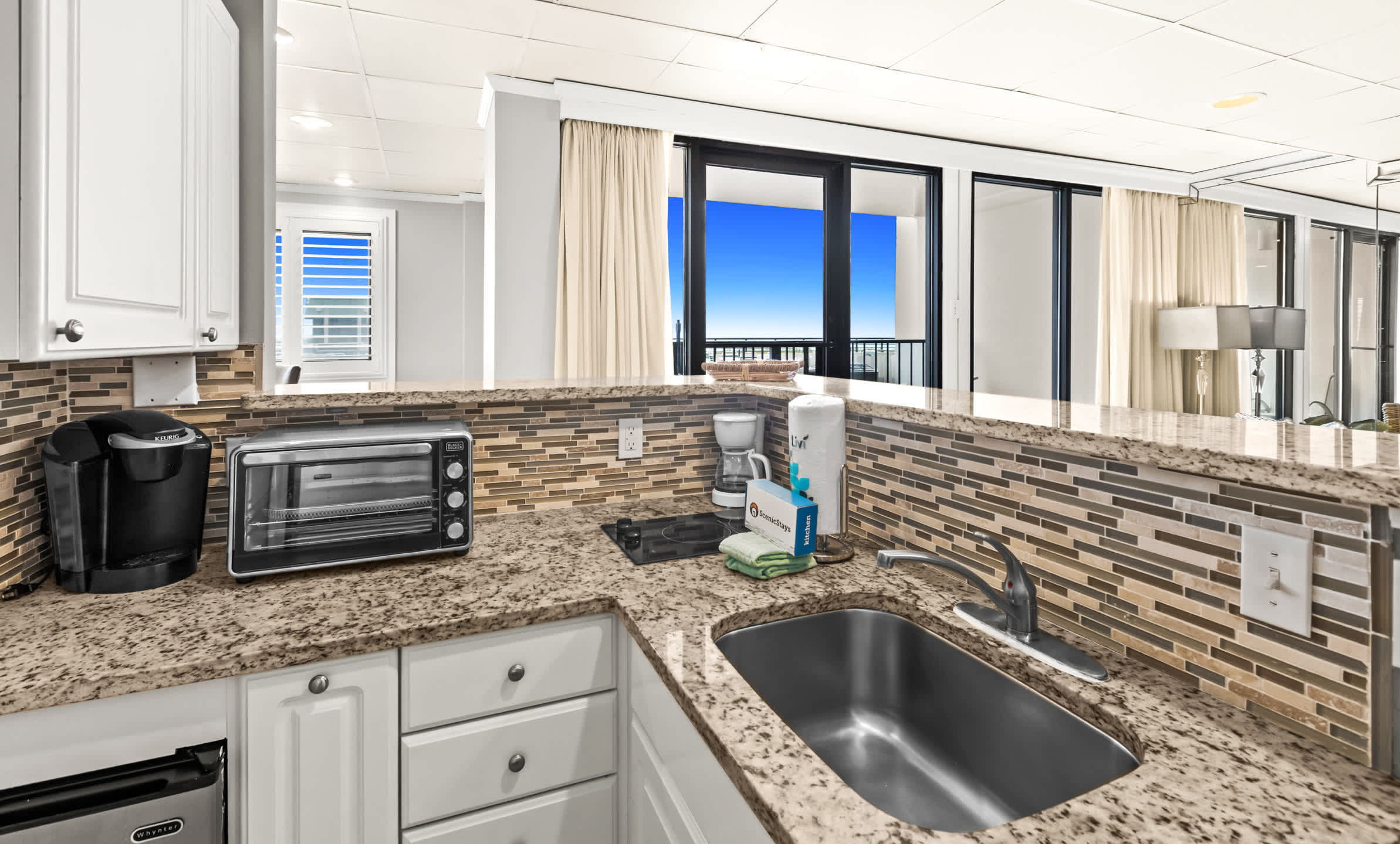 A close-up view of the kitchenette counter with granite countertops and a mosaic tile backsplash, showing a coffee maker, toaster oven, and a small stovetop, with a glimpse of the balcony and ocean through sliding glass doors.