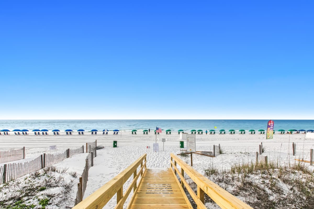 Wooden boardwalk providing direct access to a wide, white sand beach lined with rows of blue and green beach umbrellas and chairs, with the turquoise ocean stretching to the horizon under a clear blue sky.